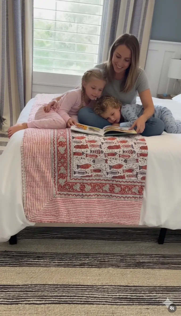 Red fish block print Kantha quilt draped over white bed, mother reading with two children in cozy bedroom setting